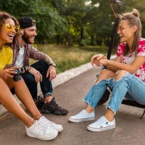 happy young company of smiling friends sitting in park on grass with electric kick scooter, man and women having fun together, colorful summer hipster fashion style, talking, smiling, making photos