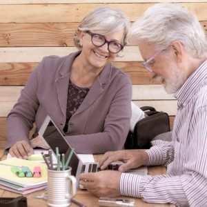 Rustic wooden office with a cheerful couple of people with white hair sitting outdoor and working with laptop and books. Camera on the background